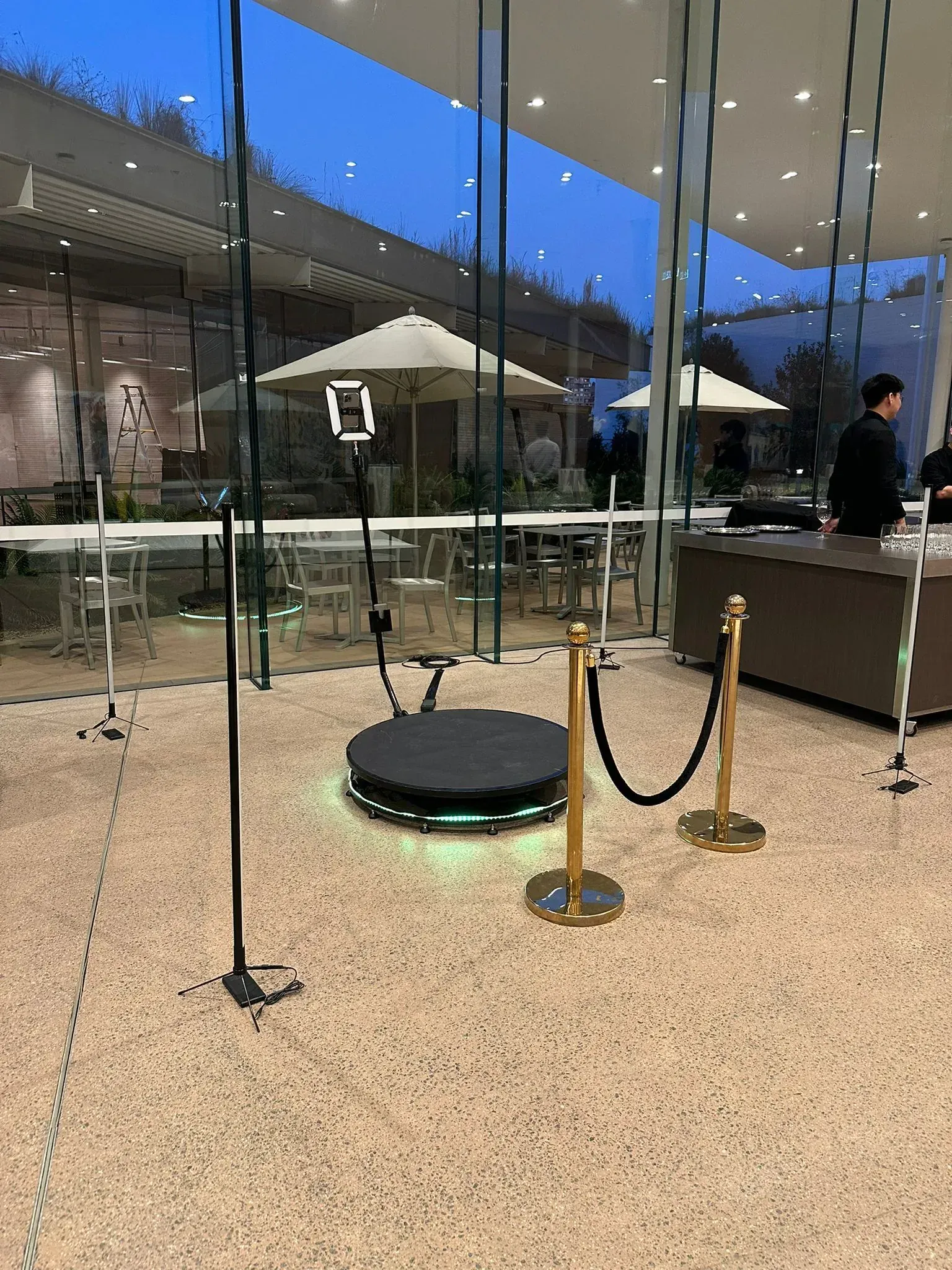 A 360 photo booth platform with ring light, set up beside gold rope bollards at an indoor venue.