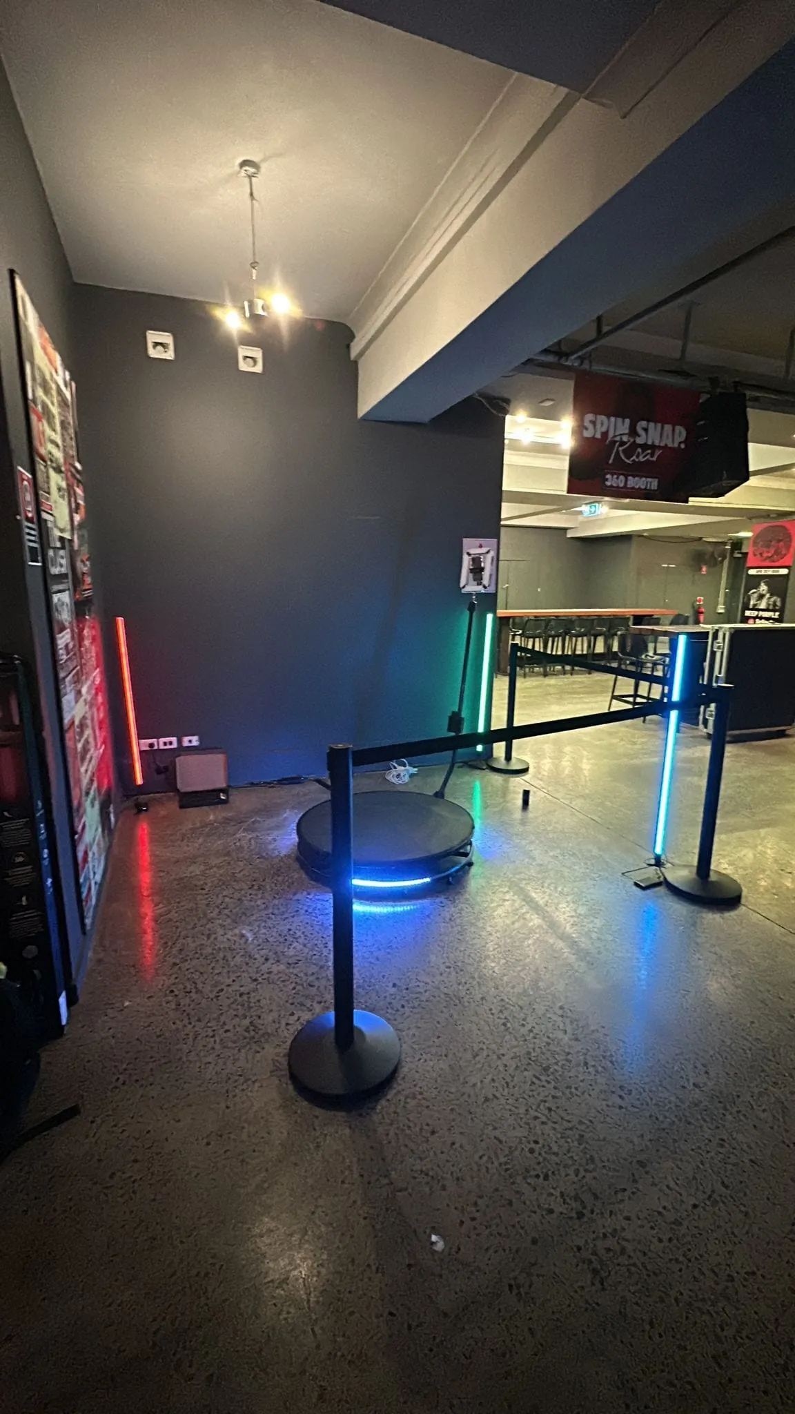 A 360 photo booth spin platform set up with LED light poles and a queue barrier inside a dimly lit venue.