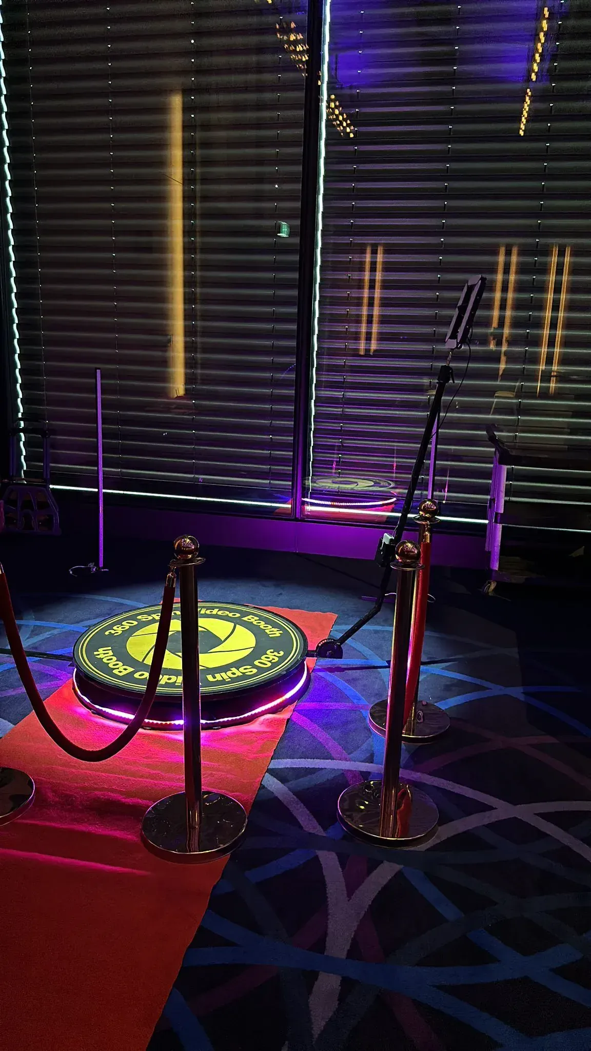 Illuminated 360 spin video booth platform on a red carpet framed by gold rope barriers in a darkened venue.