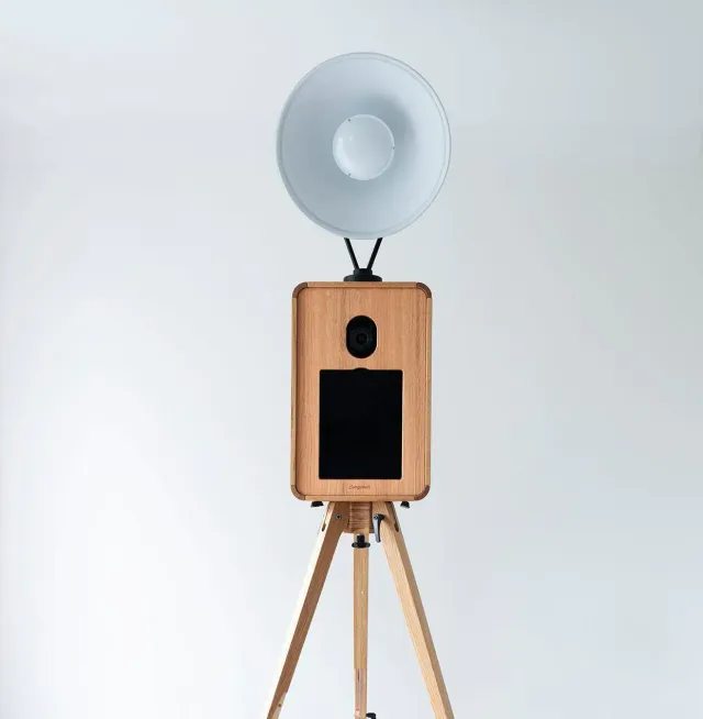 Glam photo booth: a wooden-framed booth with a large ring light on a wooden tripod, against a plain studio backdrop.