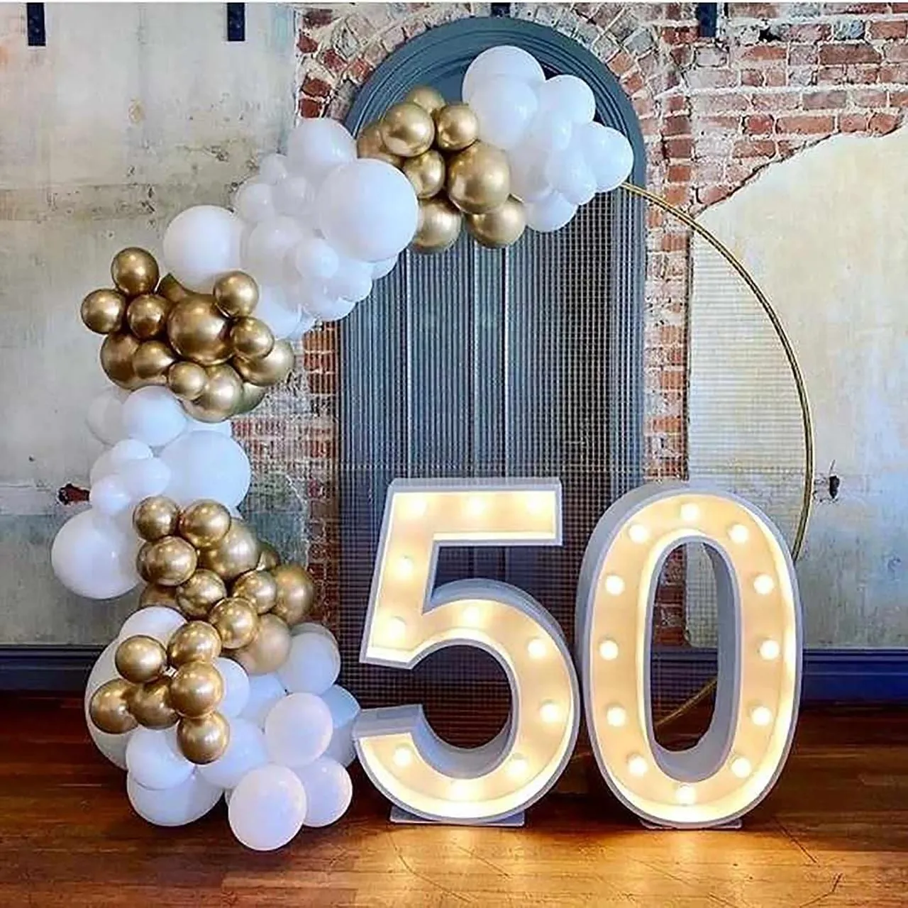 Light-up marquee number 50 beside a white and gold balloon garland against an exposed-brick wall.