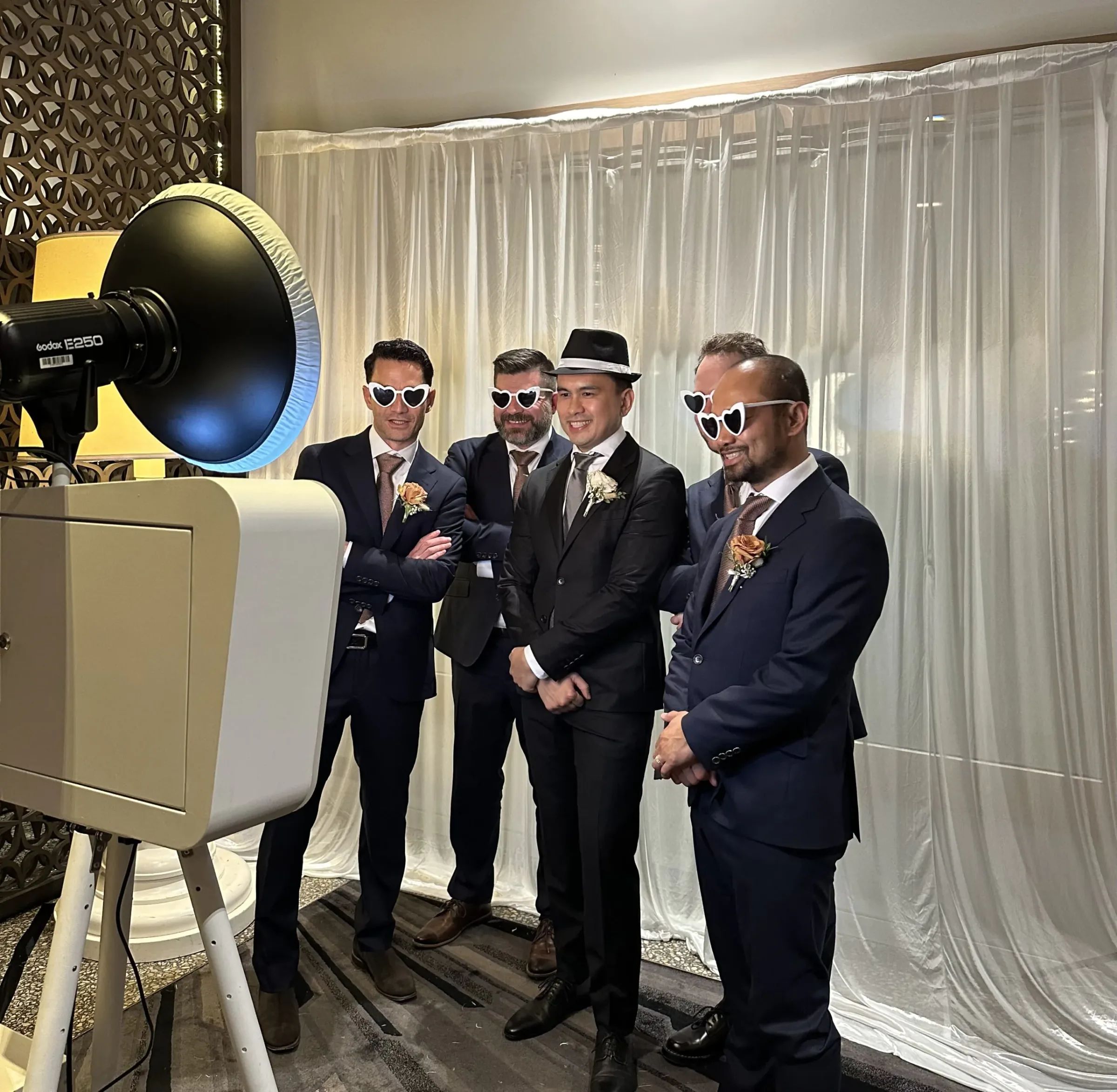 Five men in suits wearing heart-shaped sunglasses pose beside an open-air photo booth in front of a sheer curtain backdrop at a wedding.