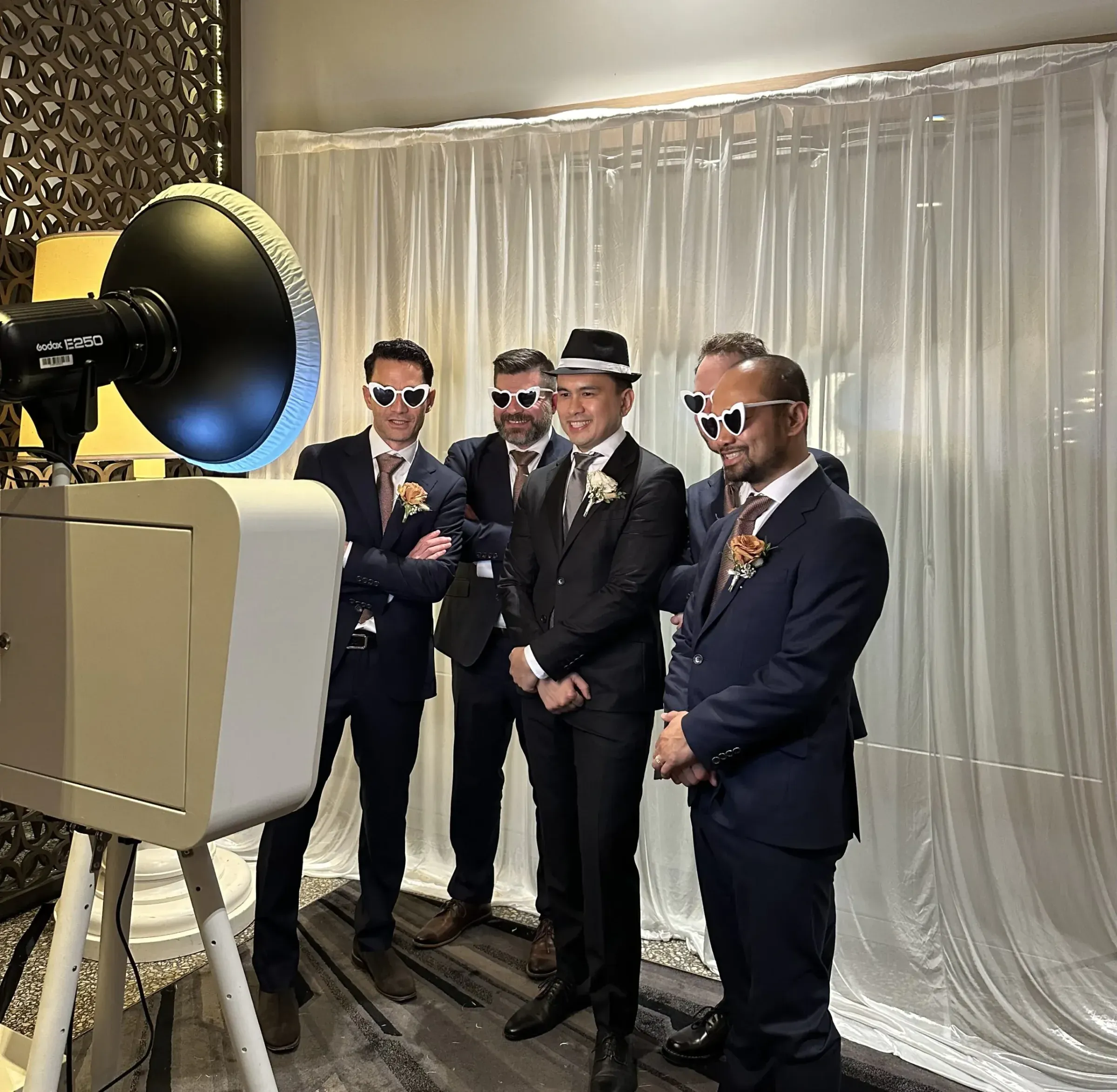 Five men in suits wearing heart-shaped sunglasses pose beside an open-air photo booth in front of a sheer curtain backdrop at a wedding.