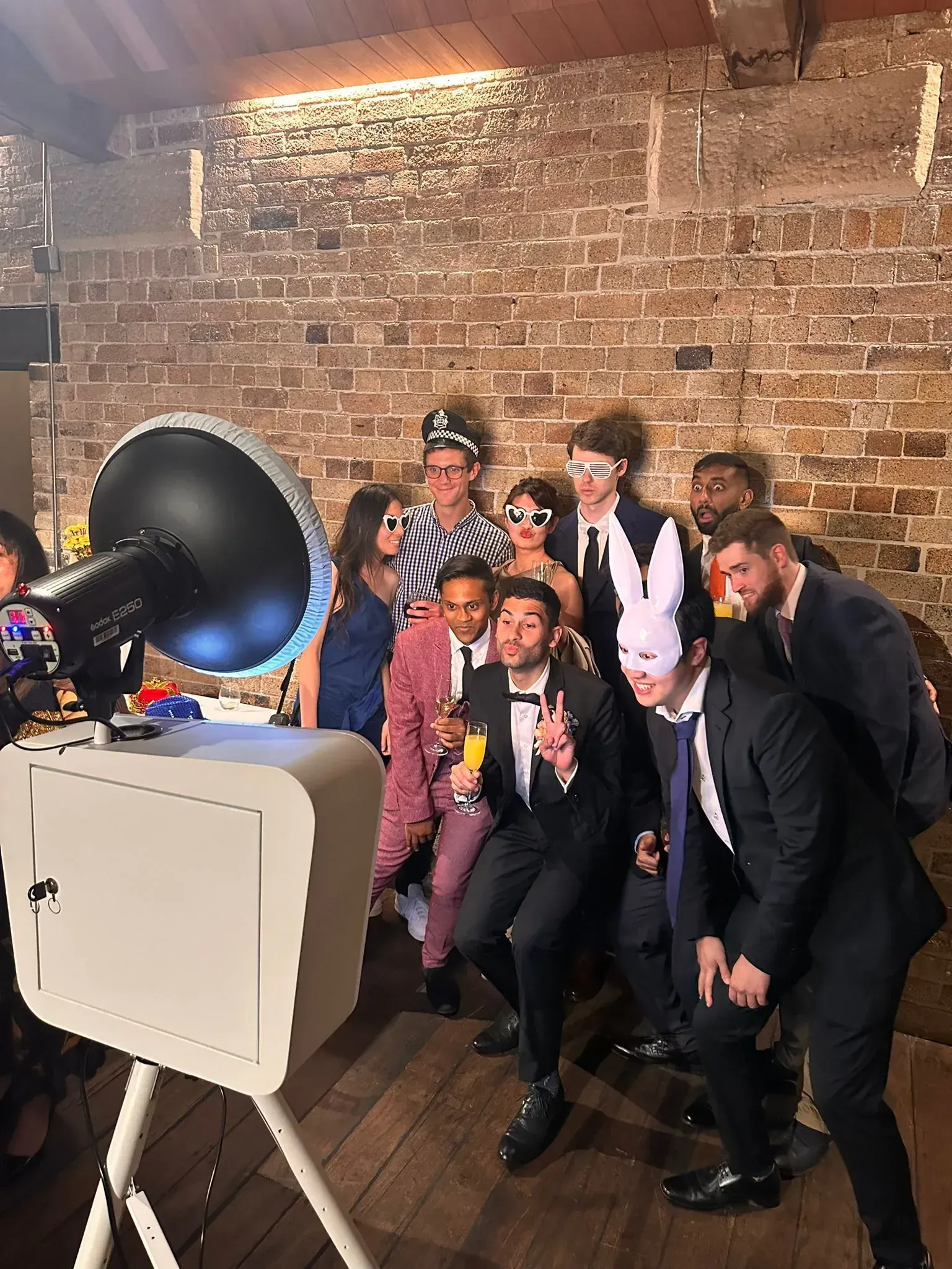 A group of guests in suits and dresses posing with a bunny mask, novelty glasses and other props beside an open-air photo booth against an exposed brick wall.