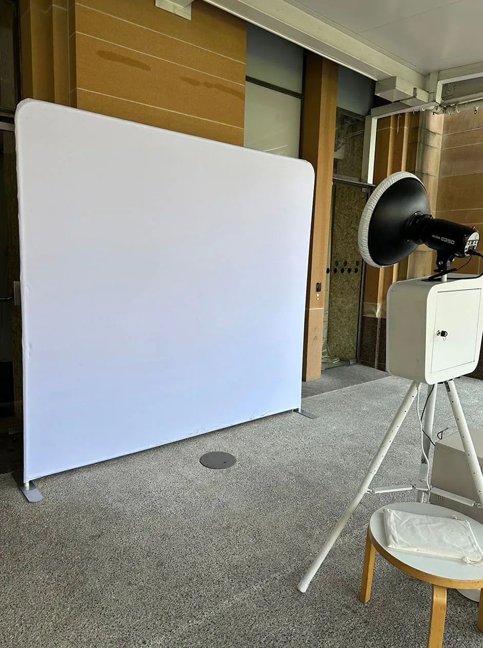 Open-air photo booth with a ring flash set up in front of a plain white backdrop on a polished concrete floor.
