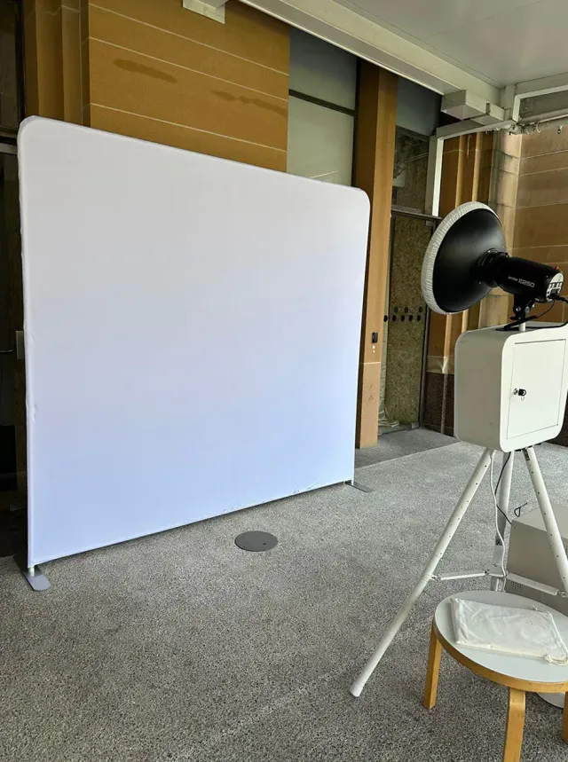 Open-air photo booth with a ring flash set up in front of a plain white backdrop on a polished concrete floor.