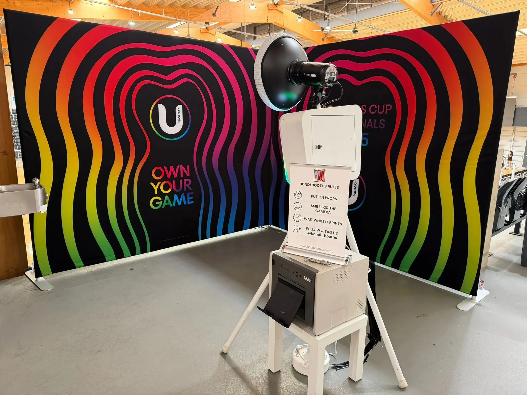 White open-air photo booth on a tripod with a beauty-dish light and print instructions sign, set up in front of a colourful rainbow backdrop indoors.