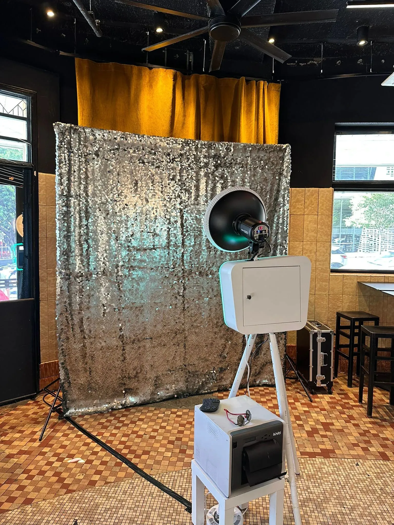 White open-air photo booth on a tripod set up in front of a silver sequin backdrop inside a tiled venue.