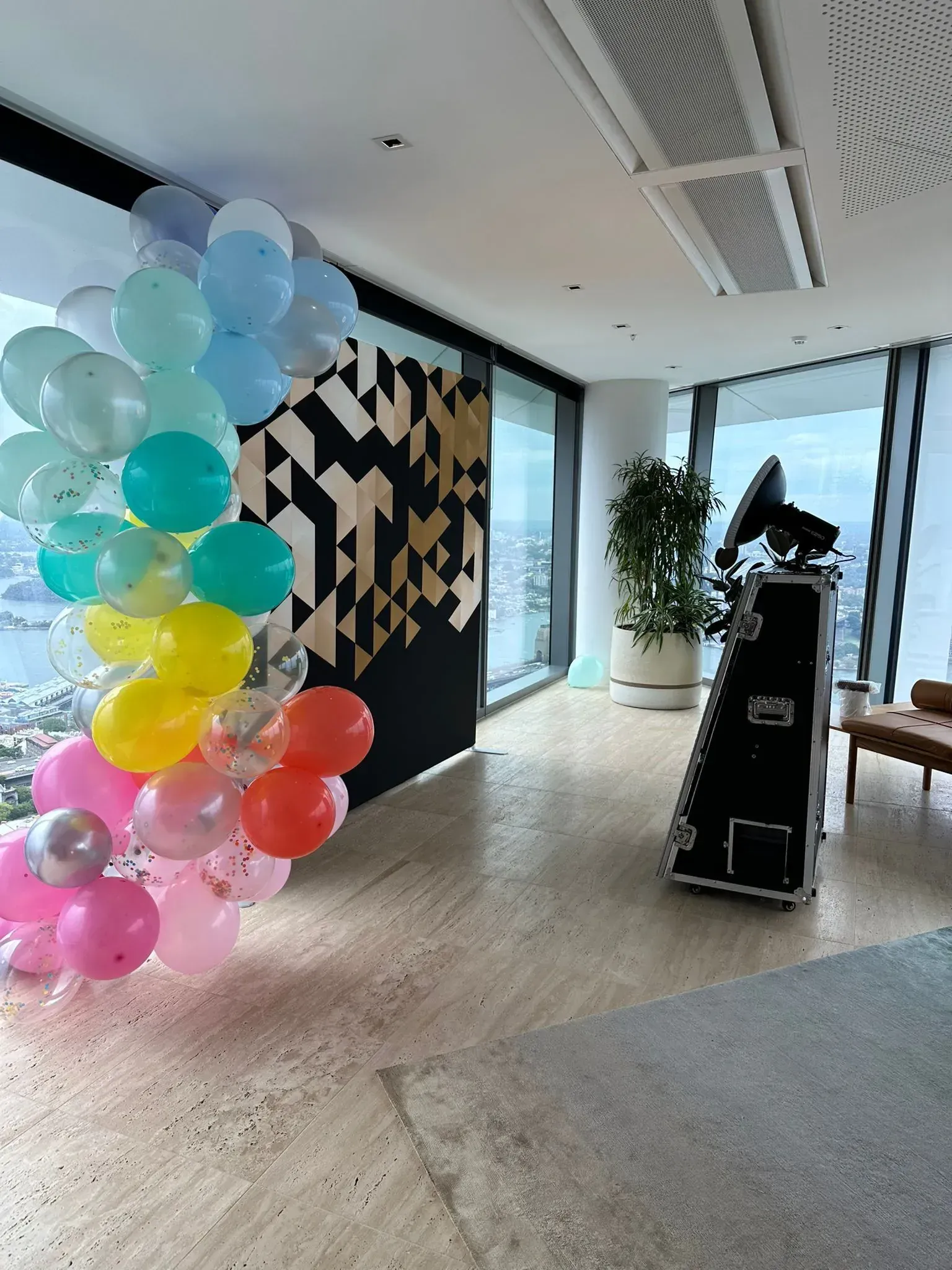 Photo booth beside a pastel balloon garland in a high-rise office with harbour views through floor-to-ceiling windows.