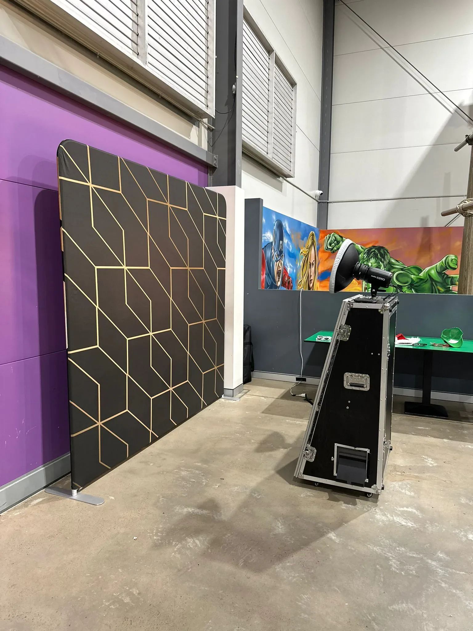 Photo booth with a black and gold geometric backdrop and flight-case booth set up in a warehouse space with a superhero mural.