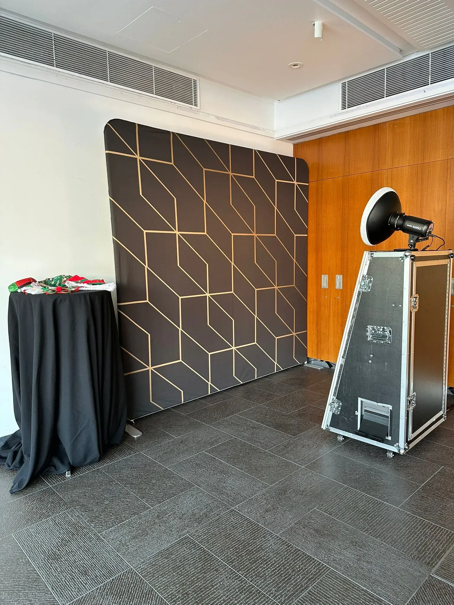 Photo booth setup with a black and gold geometric backdrop and a flight-case booth with a beauty-dish light in an event room.