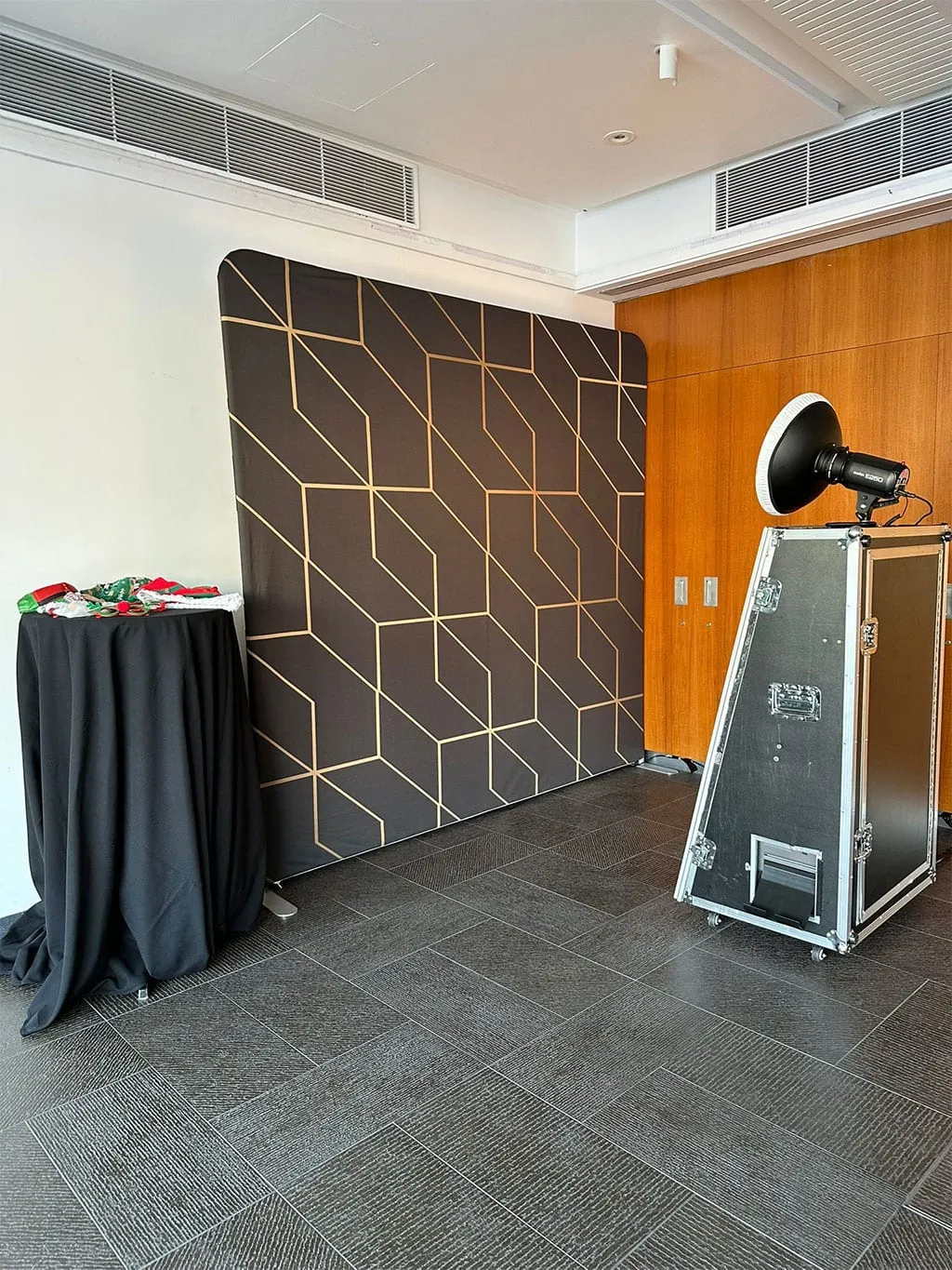 A black and gold geometric backdrop set up next to booth lighting and a flight case on a tiled floor