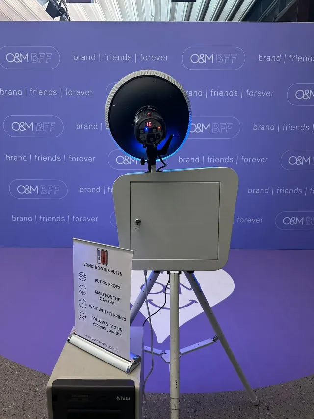 A Bondi Booths photo booth on a tripod in front of a purple branded media wall, with a printer and a booth rules sign alongside
