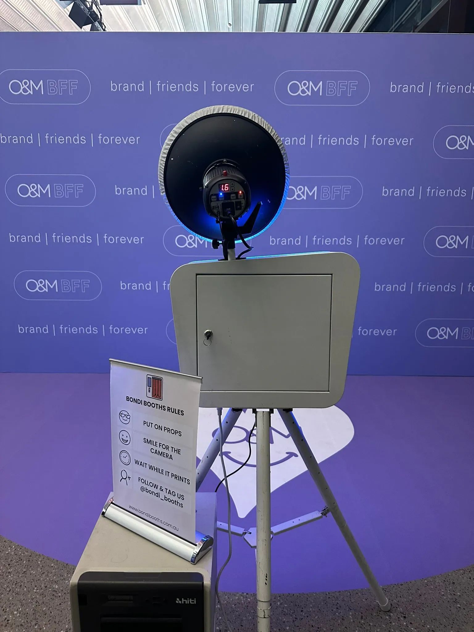 A Bondi Booths photo booth on a tripod in front of a purple branded media wall, with a printer and a booth rules sign alongside