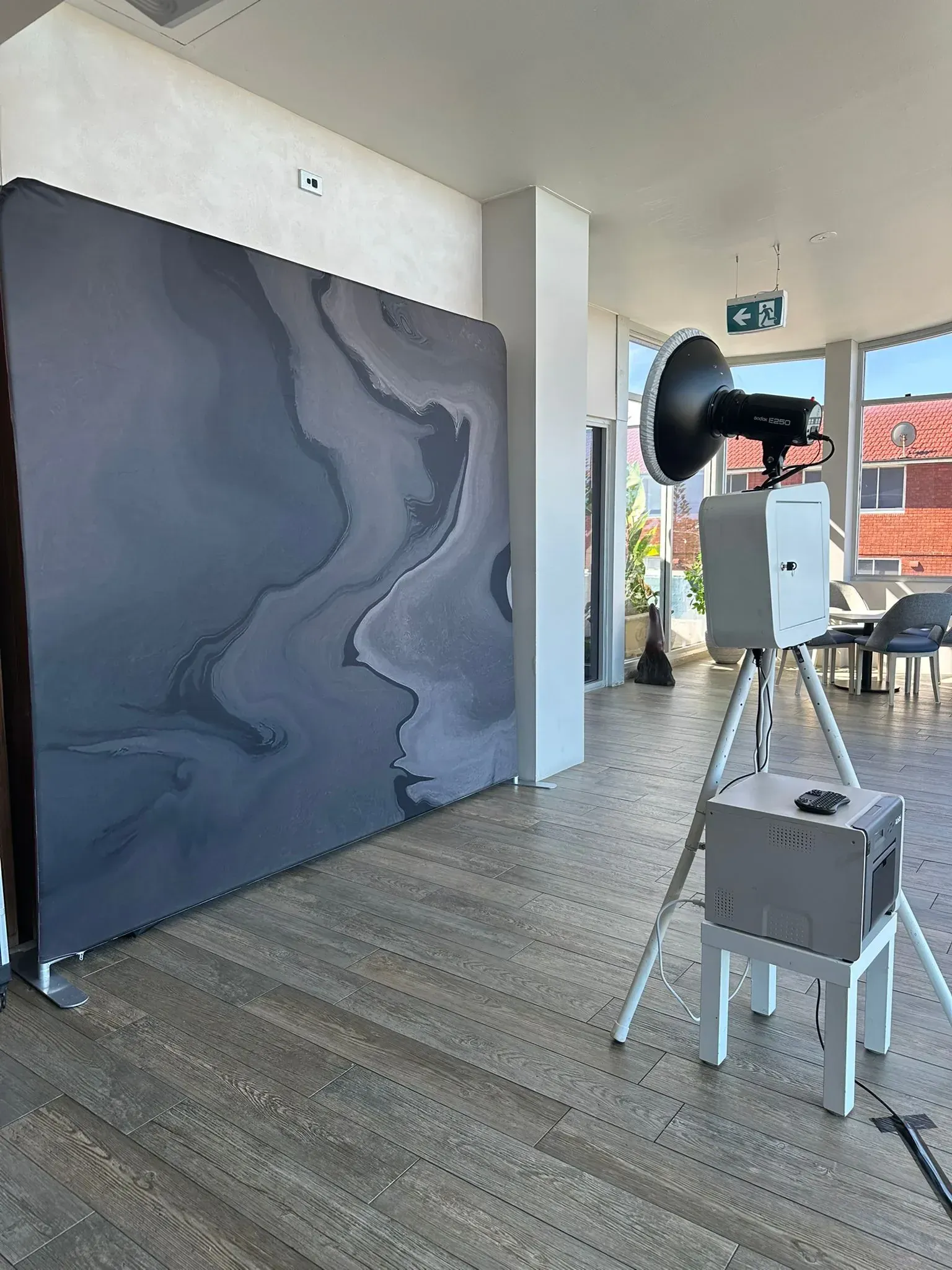 Photo booth on a tripod with a grey marble backdrop set up in a bright room with windows overlooking rooftops.
