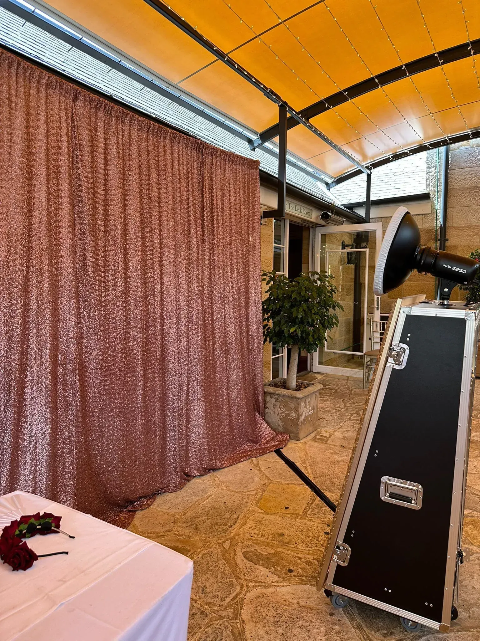 Photo booth with a rose gold sequin backdrop set up in a covered courtyard beside a potted tree and a rose-topped table.