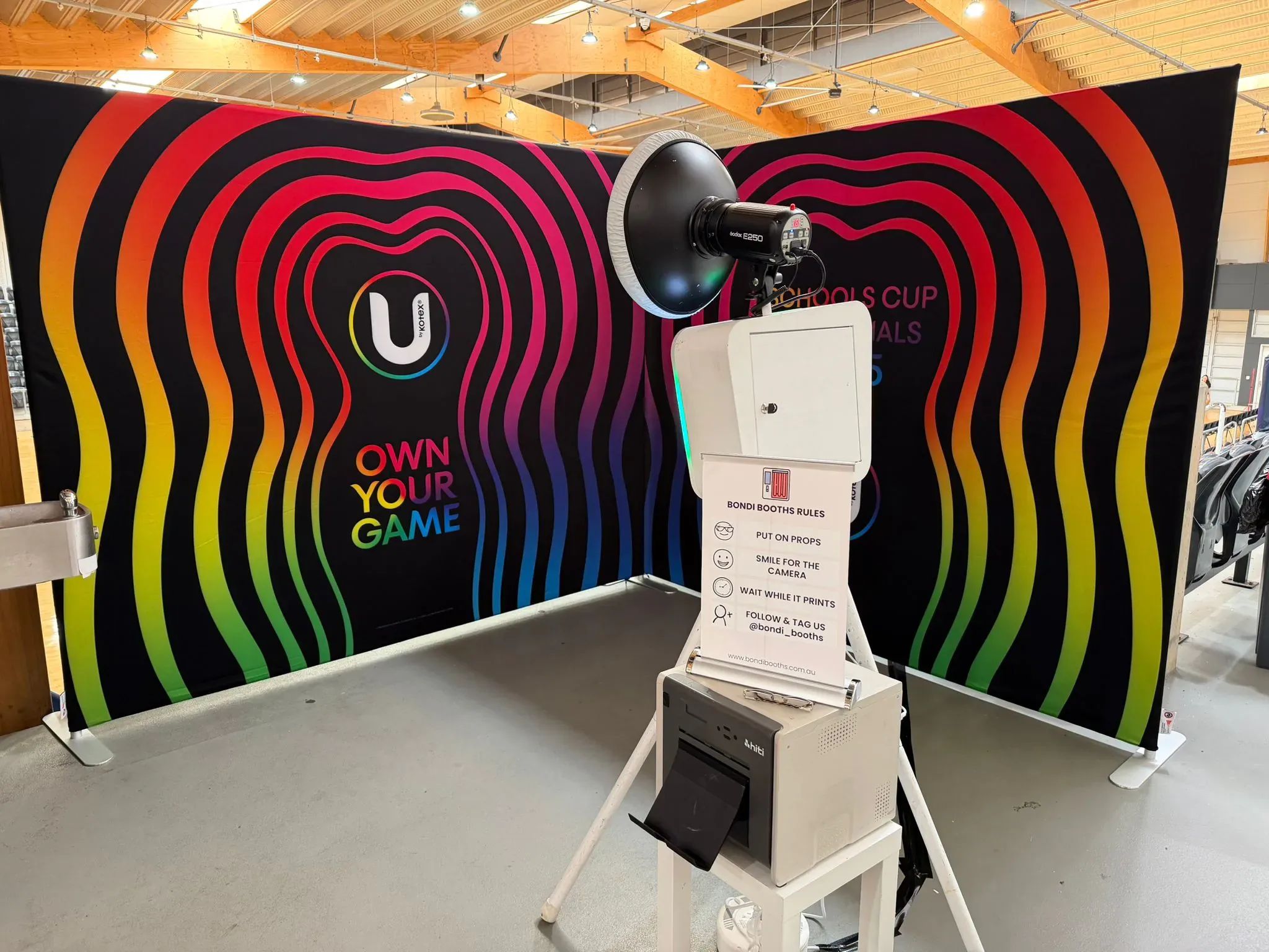 Photo booth on a tripod with a printed rainbow Own Your Game backdrop and a Bondi Booths rules sign at a sports event.