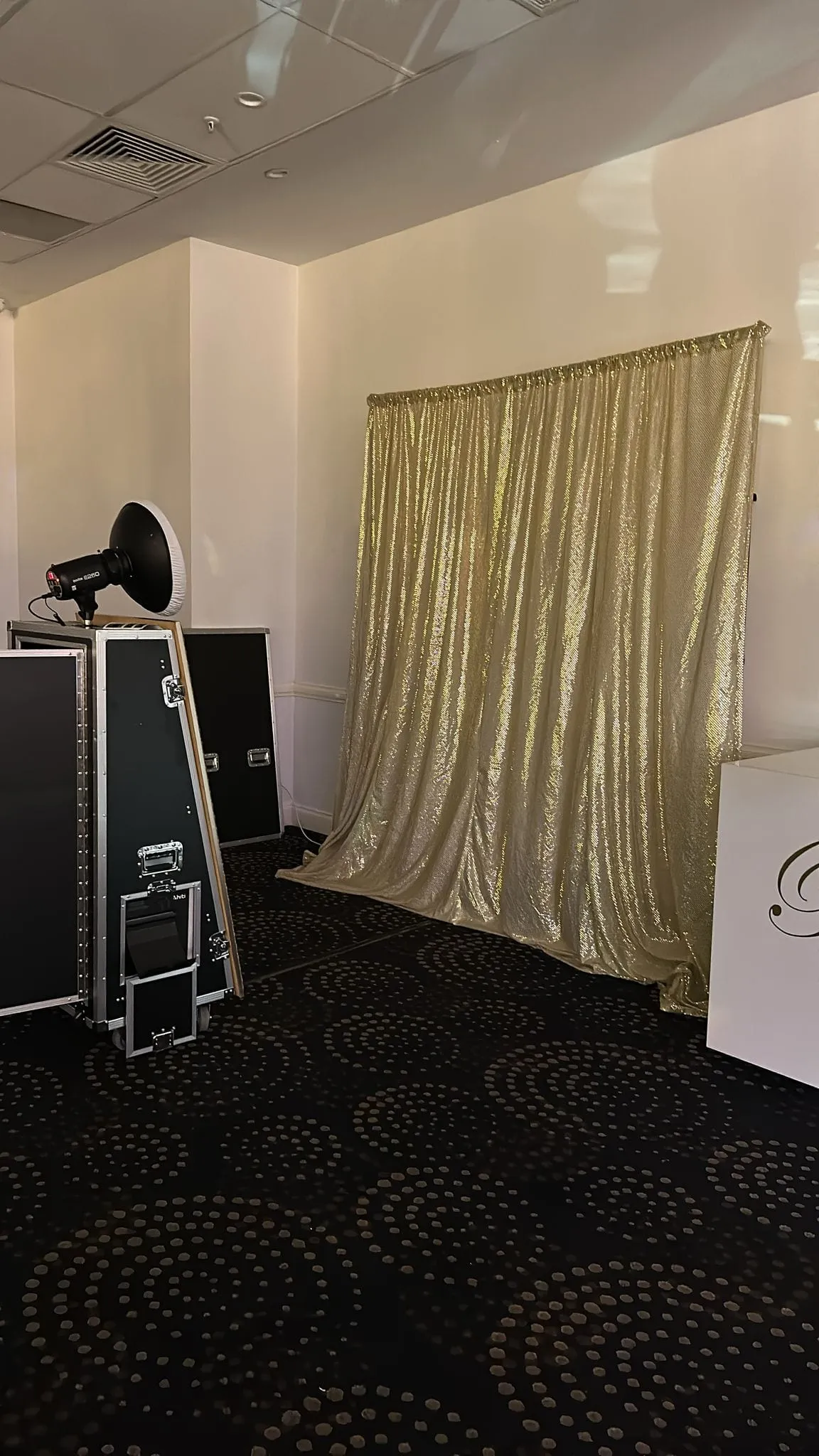 A photo booth set up beside a gold sequin backdrop and flight cases in a carpeted function room