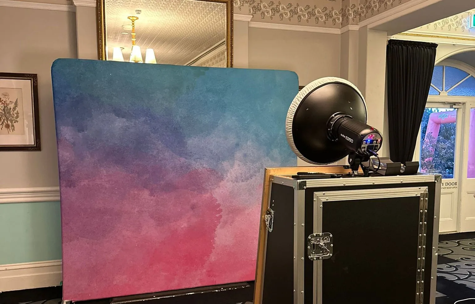 A pink and blue watercolour backdrop set up beside booth lighting and a flight case in a heritage function room