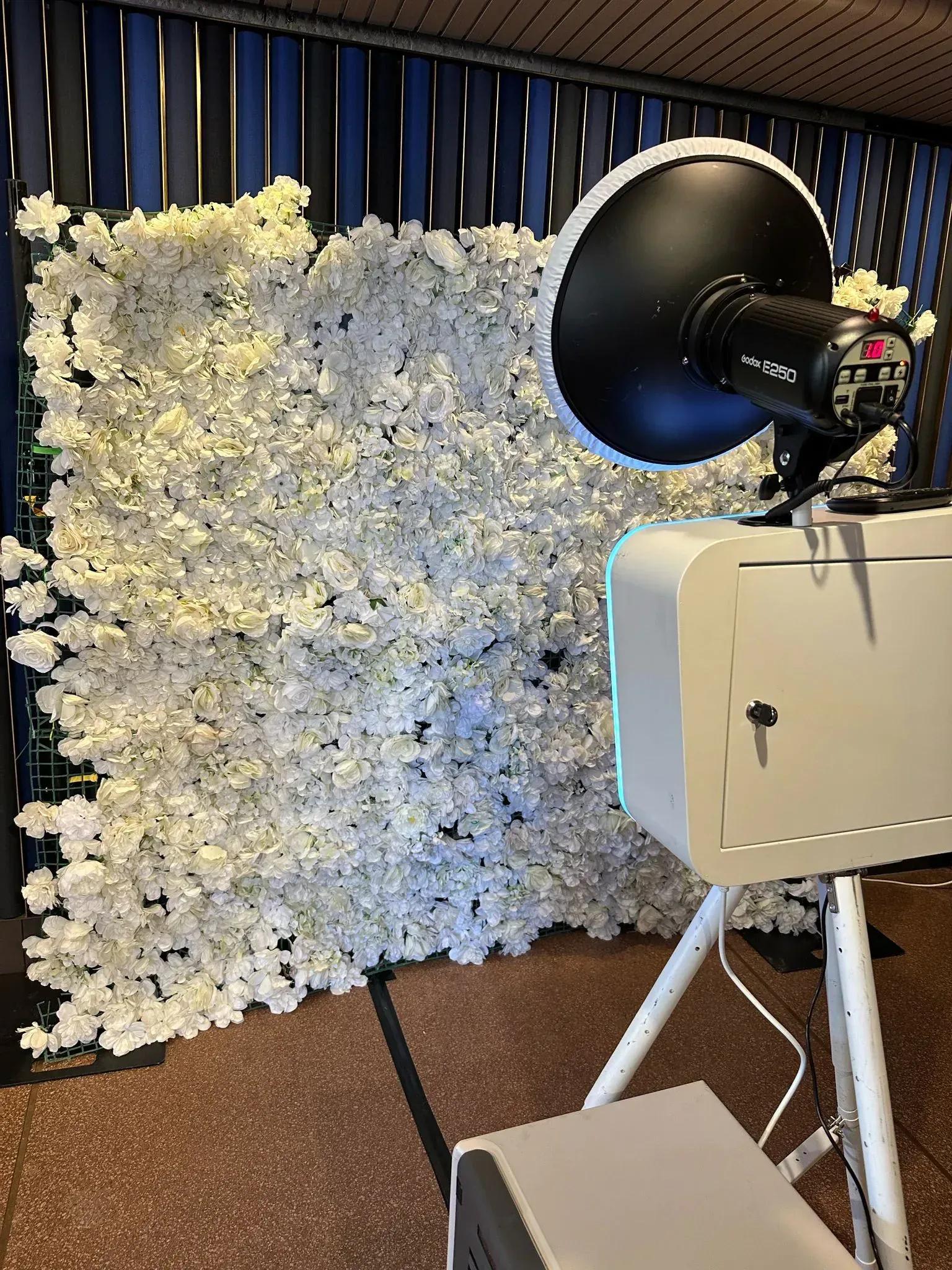 A photo booth on a tripod with a beauty-dish flash in front of a white flower wall backdrop